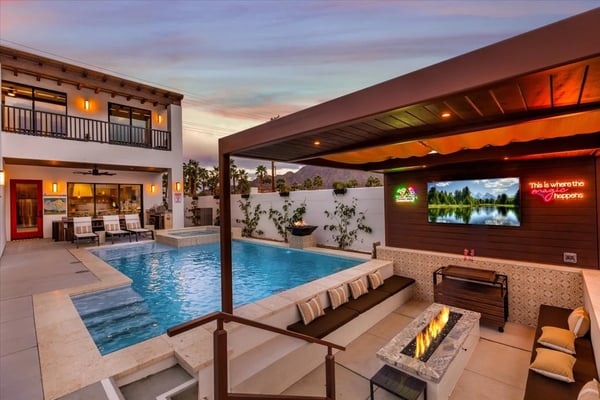 Backyard with illuminated swimming pool, lounge seating, fire pit, covered patio with TV, and mountain views at dusk