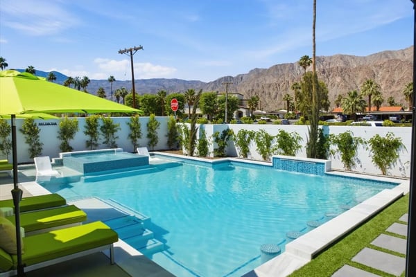 Resort swimming pool with turquoise water, white loungers, lime-green shade sail, and desert mountains in background