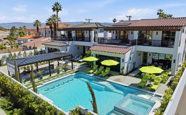 Aerial view of modern resort with turquoise pool, lime-green lounge chairs, white buildings with terracotta roofs, and palm trees.